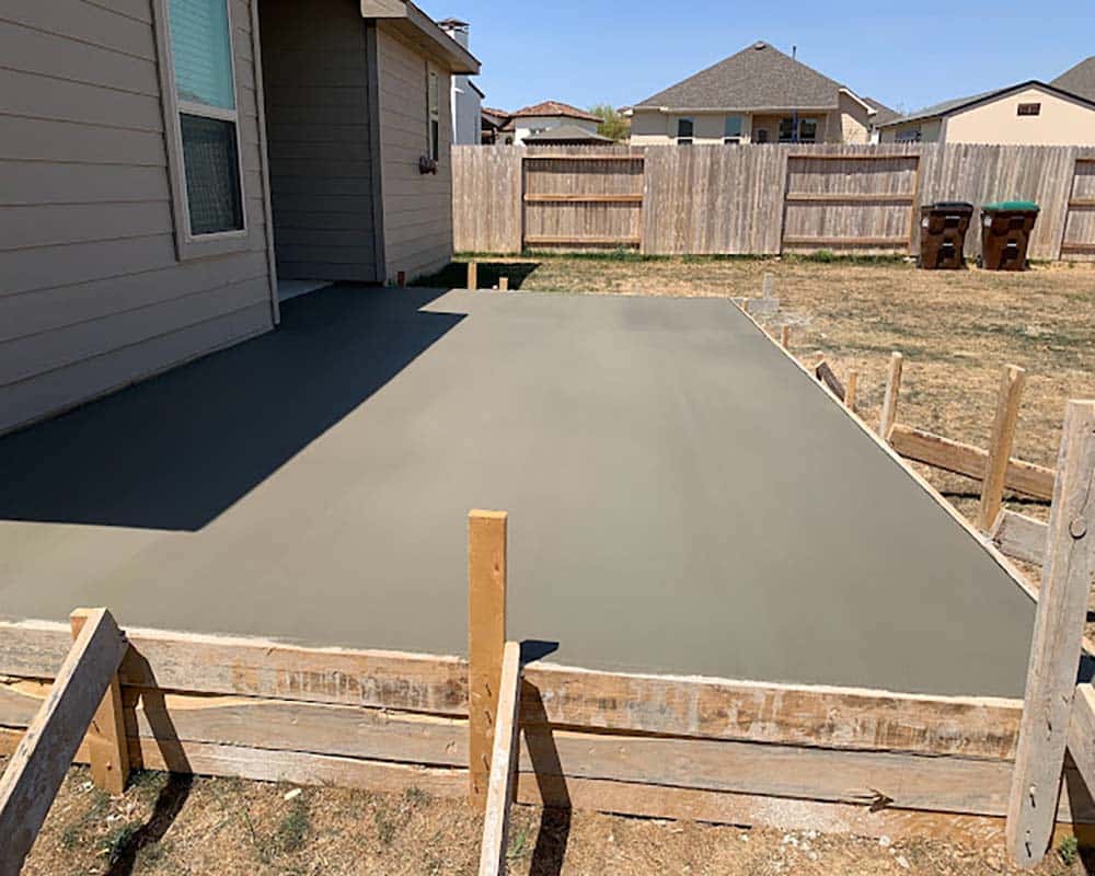 Freshly poured smooth concrete patio in a backyard with wooden formwork, adjacent to a gray house and wooden fence.