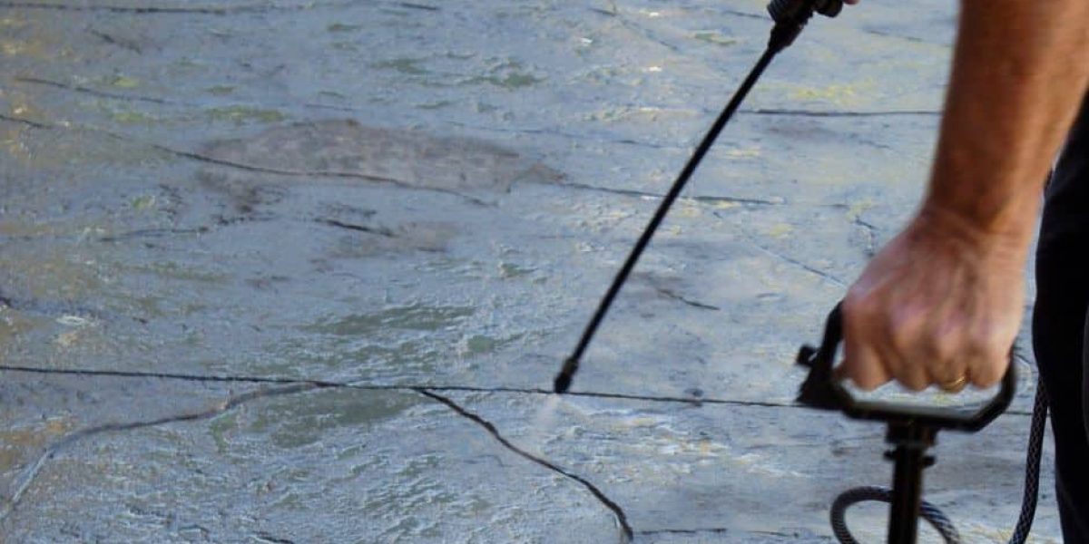 Person spraying sealant on textured concrete driveway.