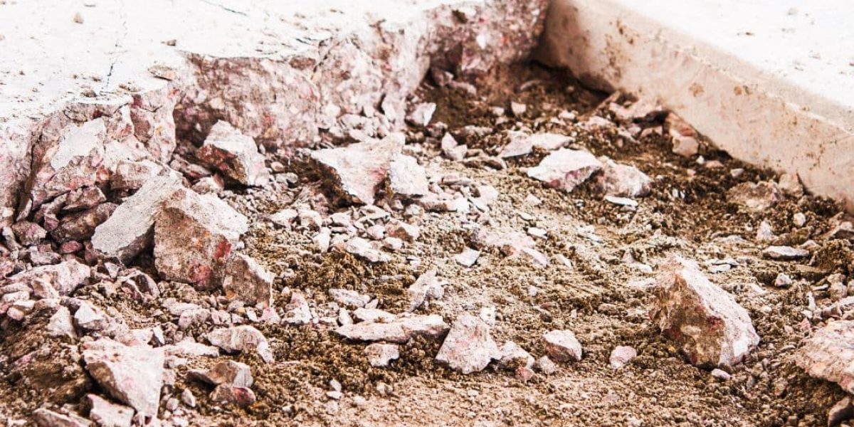 The image shows a close-up of a demolished concrete edge next to an intact concrete surface. There are scattered chunks of broken concrete and rubble on the ground, indicating recent construction work or demolition. The textures of the concrete pieces and the dirt ground are clearly visible, suggesting the site is under active construction or renovation.