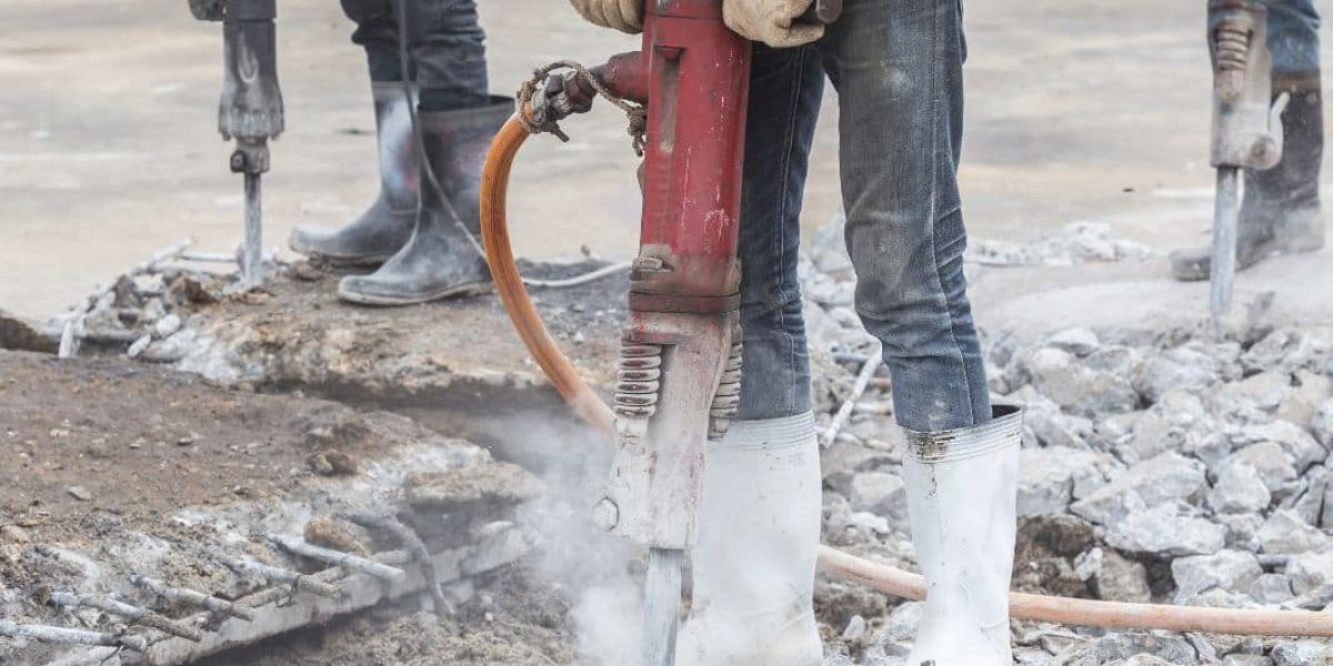 The image depicts a construction worker operating a jackhammer on a concrete surface. The worker is wearing safety boots, jeans, and a red shirt, and is holding the jackhammer which is actively breaking up the concrete, evidenced by the visible dust and debris. There's another jackhammer in the background, suggesting a team is working on demolishing or renovating the area. Safety and protective gear appear to be a priority on this site.