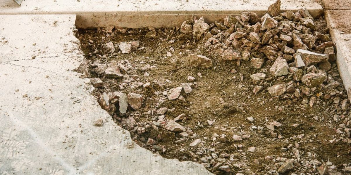 The image shows a close-up view of a construction or repair site. There is a section where the concrete has been removed or has crumbled away, revealing the earth and broken pieces of concrete below. Around the edges, intact concrete slabs can be seen, and in the background, there is a pile of small stones, indicating recent work on the surface. A yellow tool, possibly a level or a measuring device, is also visible, suggesting ongoing construction or repair work.