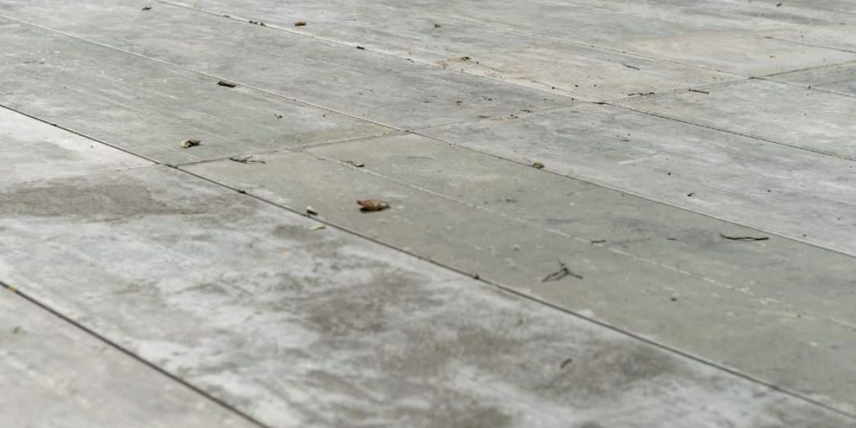 The image shows a close-up view of a concrete sidewalk or floor with defined board patterns resembling wooden planks. The surface has some dirt and small debris scattered across it, indicating recent use or exposure to the elements. The concrete has a matte finish, with slight variations in color and texture giving it a somewhat weathered appearance.