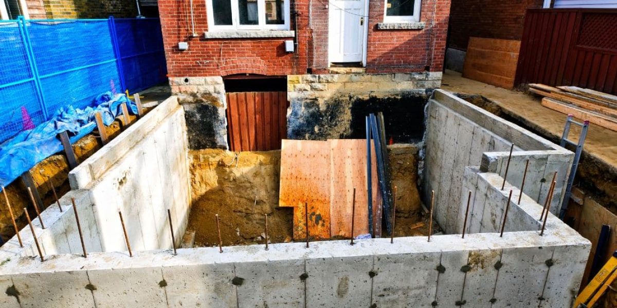 The image shows an overhead view of a construction site with a new concrete foundation in the ground adjacent to a red brick house with white windows and a wooden brown door. There is blue protective netting around the site and various construction materials scattered around.