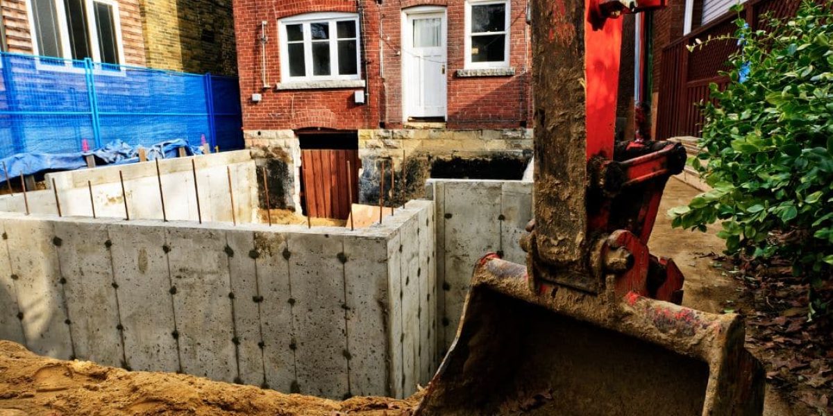 The image displays an excavator bucket near a newly constructed concrete basement or foundation in a residential area, suggesting an active construction site with a partially visible red brick house and protective blue fencing.