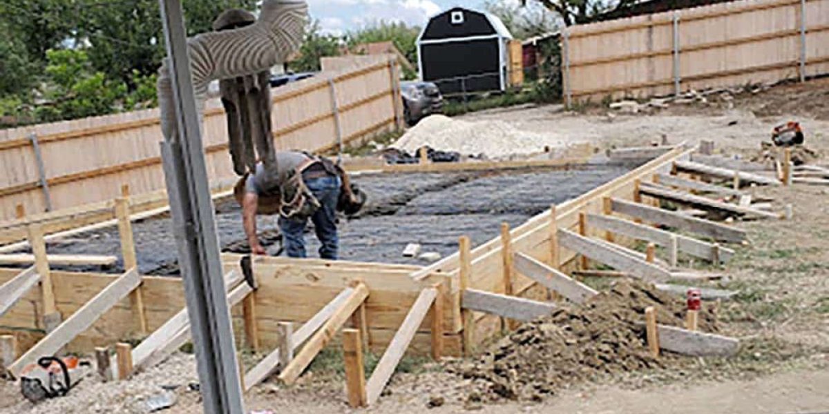 A worker levels the ground for a concrete foundation, with wooden formwork in place, in a residential backyard.