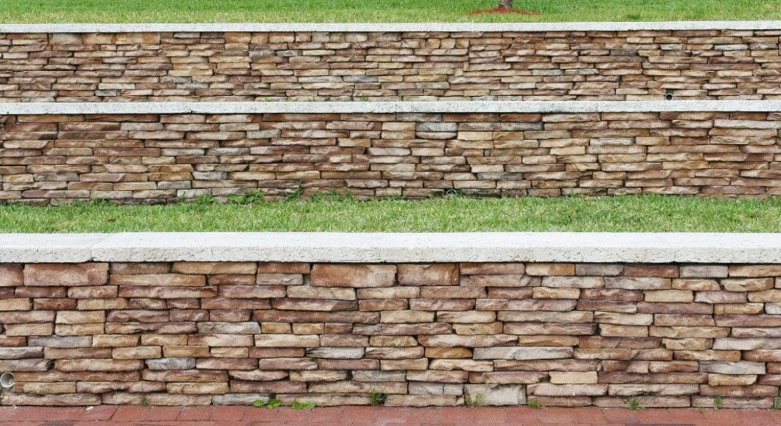 This image displays a multi-tiered retaining wall made of stacked stone in varying shades of brown and beige. The wall is set against a manicured green lawn on the top level and is adjacent to a red brick paver pathway at the bottom. The retaining wall's design is functional, helping to prevent soil erosion while providing a structured, aesthetic landscape feature. The image may represent a park or a well-maintained residential area with attention to landscape design.