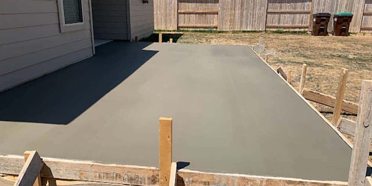 Freshly poured smooth concrete patio in a backyard with wooden formwork, adjacent to a gray house and wooden fence.