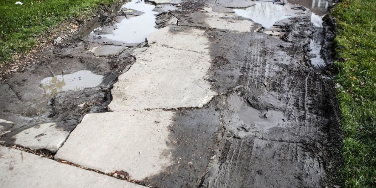 Damaged sidewalk with uneven concrete slabs and puddles.