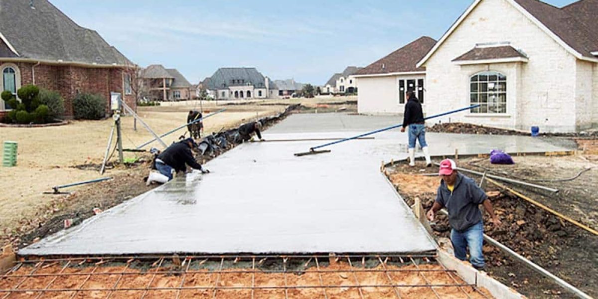 Workers pour concrete for a new driveway beside a white suburban house, with construction tools and materials visible.